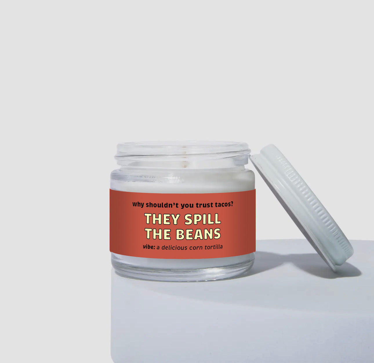 Unlit 2oz glass jar candle with a white lid, featuring a bold red label that reads: “Why shouldn’t you trust tacos? THEY SPILL THE BEANS.” Scent note on the label says: “vibe: a delicious corn tortilla.” Minimalist product photo against a light neutral background.