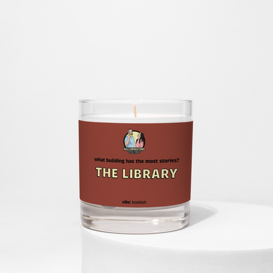 8oz coconut soy candle in a clear glass jar with a deep red label that reads “What building has the most stories? The Library.” The scent vibe is bookish. The candle is unlit and placed against a white background.