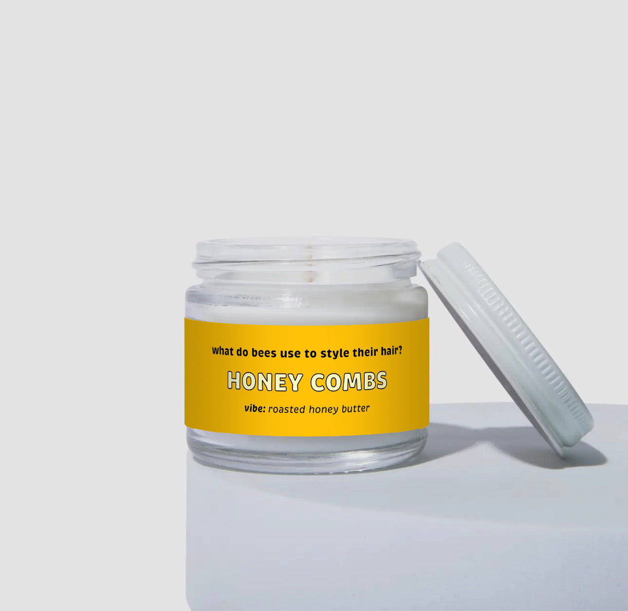 Unlit 2oz mini glass jar coconut soy candle with a white metal lid placed to the side. The candle has a bright yellow label with the text: 'what do bees use to style their hair? HONEY COMBS.' Scent description reads: 'vibe: roasted honey butter.' Clean and simple product display.