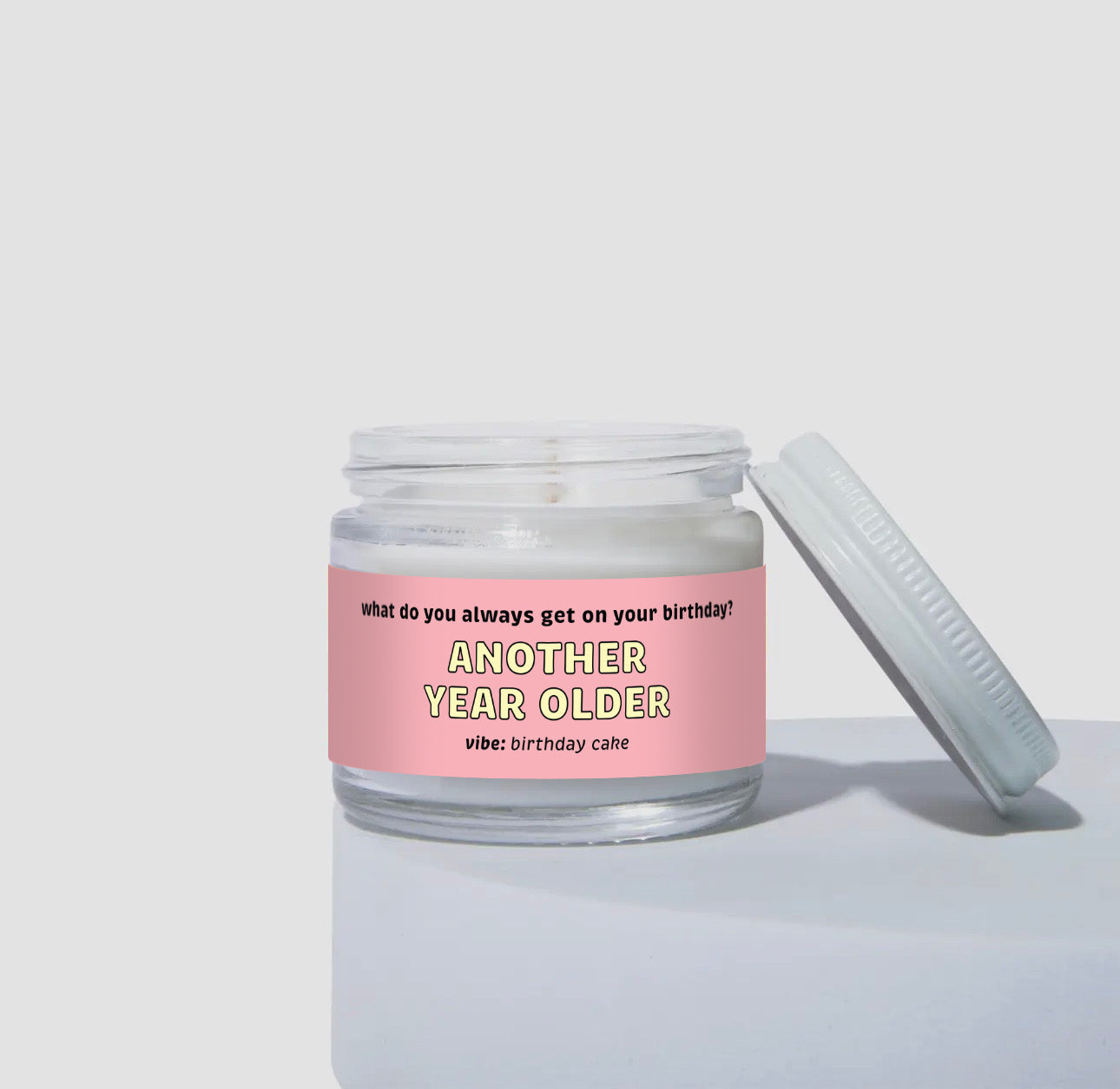 Unlit 2oz mini glass jar coconut soy candle with a white metal lid resting to the side. The candle features a light pink label that says: 'What do you always get on your birthday? ANOTHER YEAR OLDER.' Scent note reads: 'vibe: birthday cake.' Clean, minimalist product presentation.