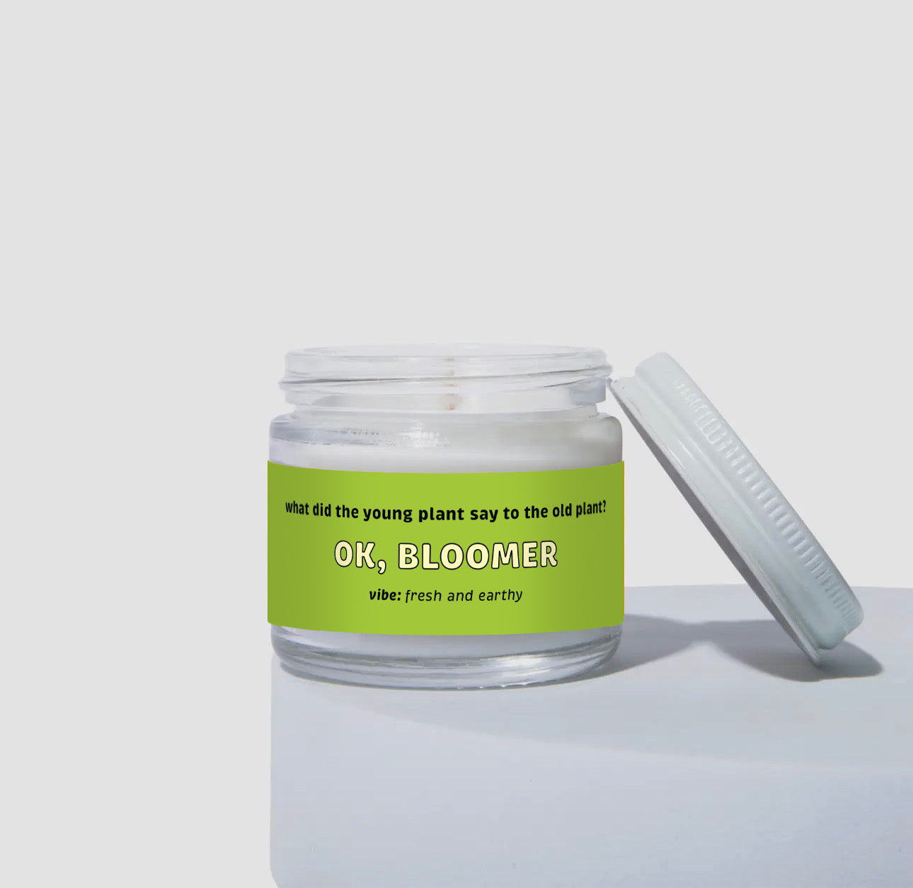 Unlit 2oz mini glass jar coconut soy candle with a white metal lid resting to the side. The candle features a bright green label that says: 'What did the young plant say to the old plant? OK, BLOOMER.' Scent note reads: 'vibe: fresh and earthy.' Clean, minimalist product presentation.