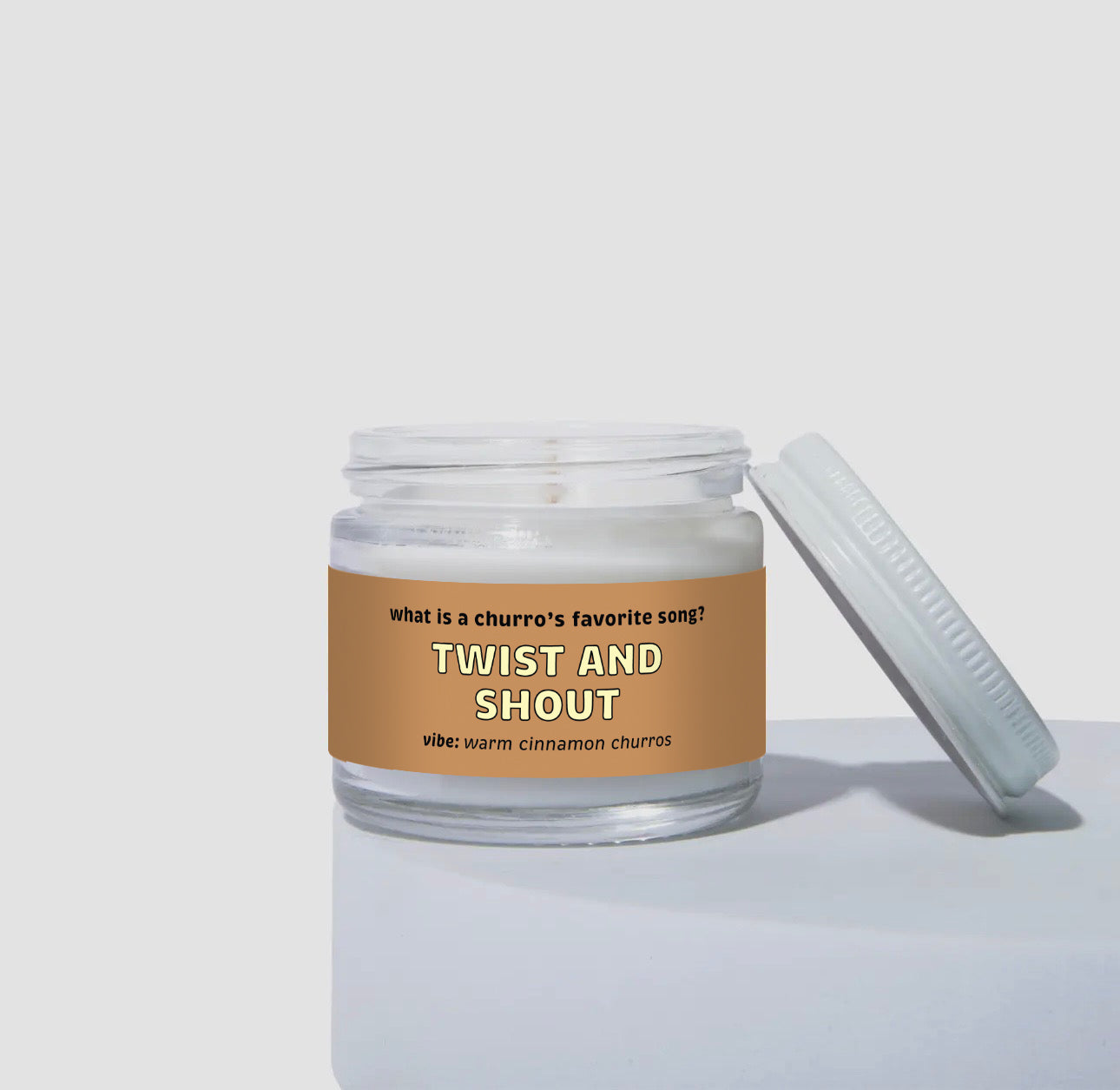 2 oz mini coconut soy candle in a clear glass jar with a white lid, labeled “Twist and Shout.” Tan-brown label with the text “What is a churro’s favorite song? Twist and Shout,” with the vibe described as “warm cinnamon churros.