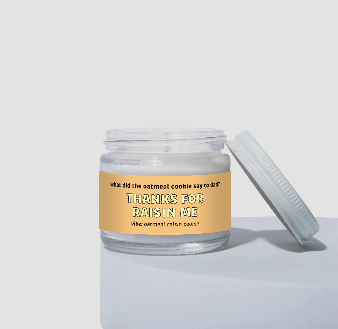 2 oz mini coconut soy candle in a clear glass jar with a light tan label that reads “Thanks for Raisin Me.” The design includes the text “what did the oatmeal cookie say to dad?” and the vibe is oatmeal raisin cookie. White wax inside jar with lid leaning against the side.