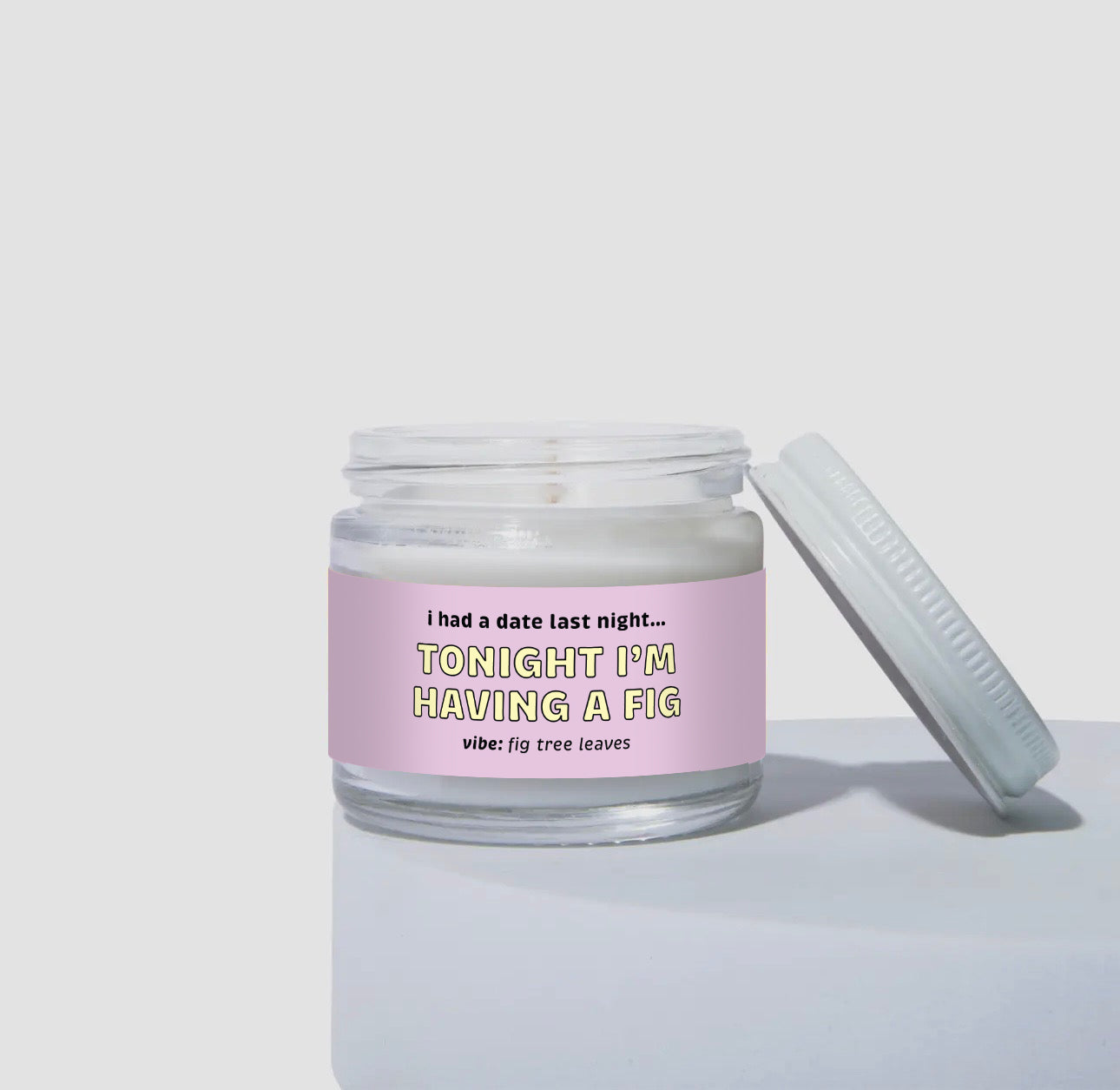 Unlit 2oz mini glass jar coconut soy candle with a white metal lid placed to the side. The candle has a light lavender label with the text: 'i had a date last night… TONIGHT I’M HAVING A FIG.' Scent description reads: 'vibe: fig tree leaves.' Clean and simple product display.