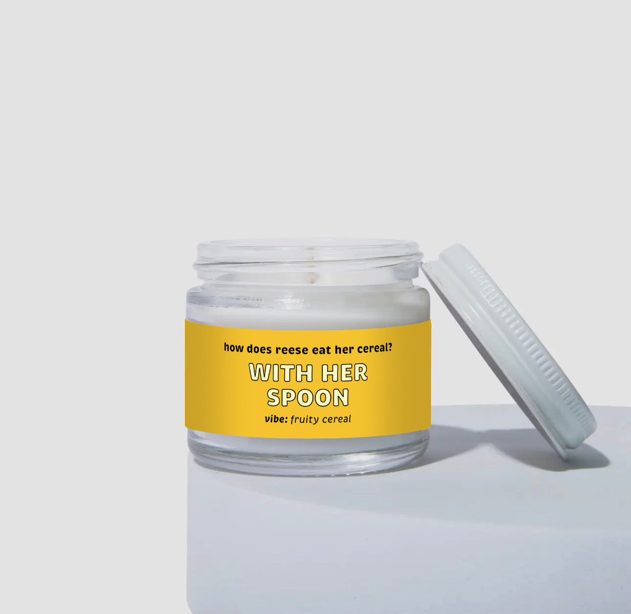 Unlit 2oz mini glass jar coconut soy candle with a white metal lid placed to the side. The candle has a bright yellow label with the text: 'how does reese eat her cereal? WITH HER SPOON.' Scent description reads: 'vibe: fruity cereal.' Clean and simple product display.