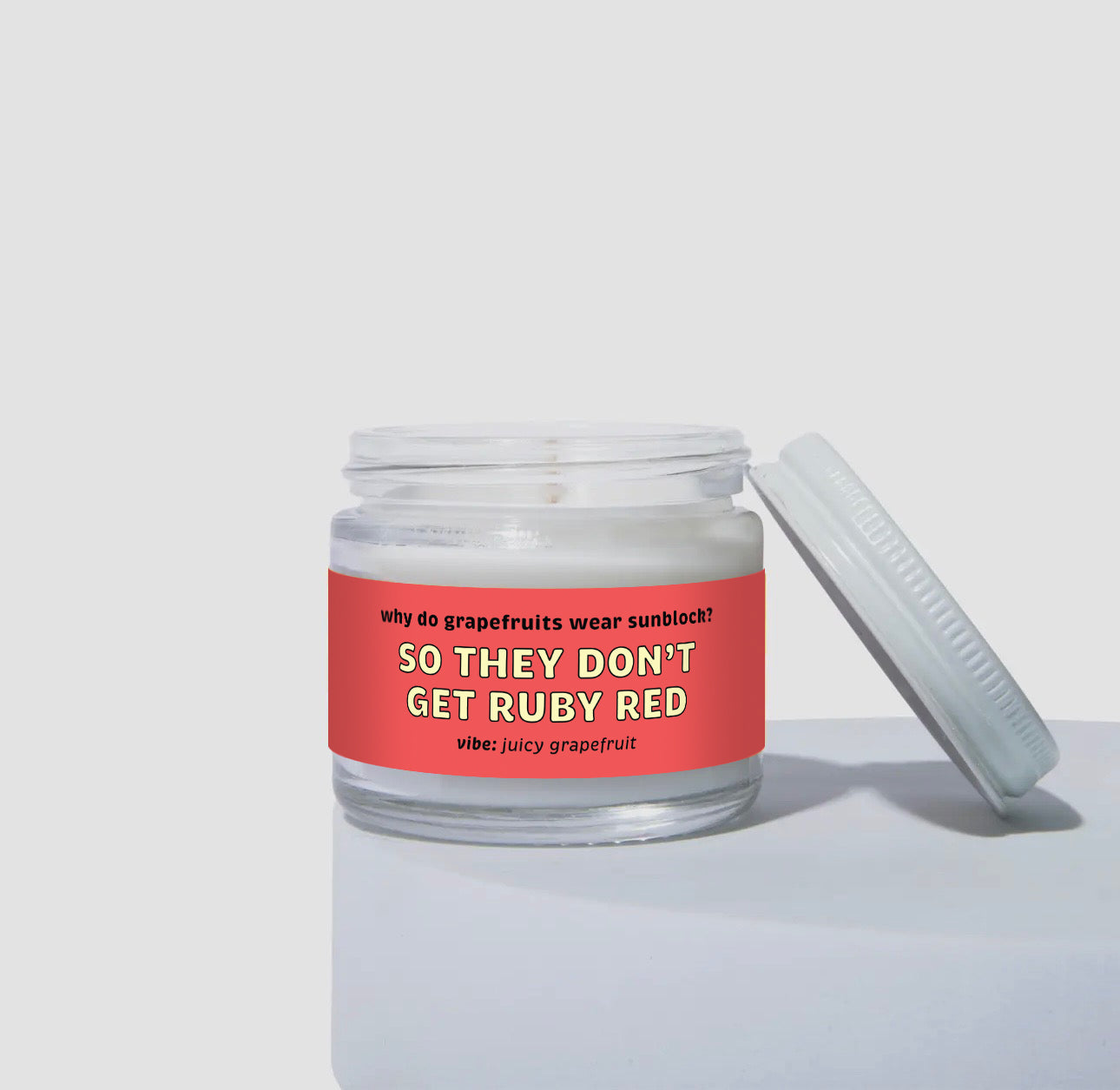 Unlit 2oz glass jar candle with a white lid, featuring a bold red label that reads: “Why do grapefruits wear sunblock? SO THEY DON’T GET RUBY RED.” Scent note on the label says: “vibe: juicy grapefruit.” Minimalist product photo against a neutral background.