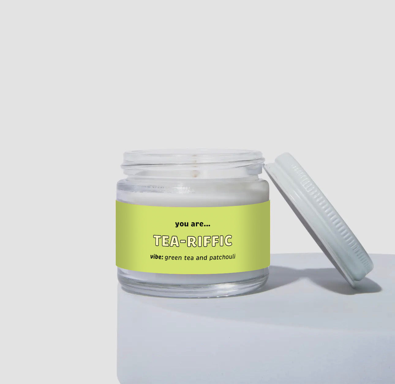 Unlit 2oz glass jar coconut soy candle with a white lid, featuring a bright green label that reads: “you are… TEA-RIFFIC.” Scent note on the label says: “vibe: green tea and patchouli.” Minimalist product photo against a light neutral background.