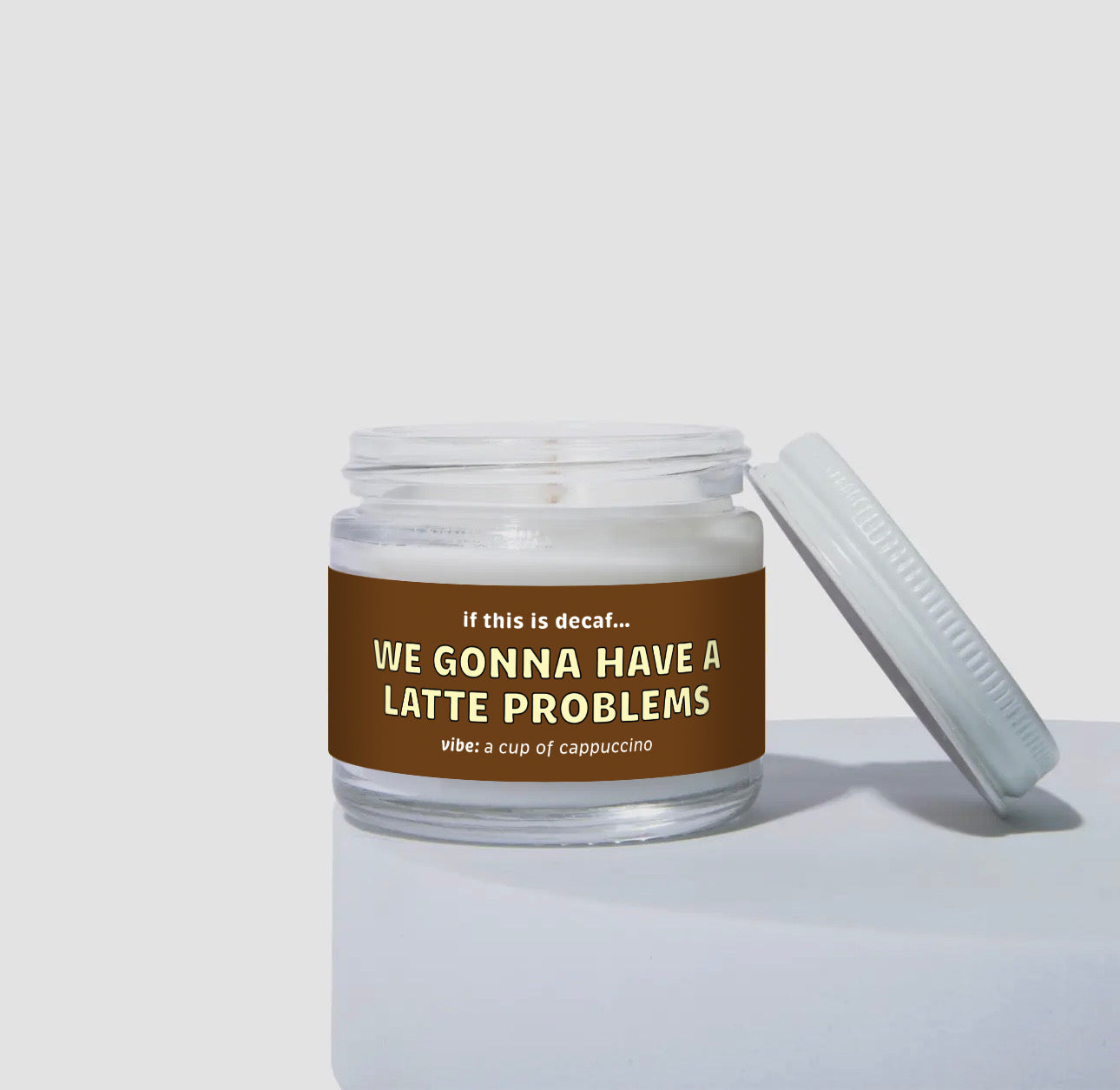Unlit 2oz mini glass jar candle with a white lid resting to the side. The candle features a rich brown label that says: 'If this is decaf… WE GONNA HAVE A LATTE PROBLEMS.' Scent description reads: 'vibe: a cup of cappuccino.' Minimalist studio product display.