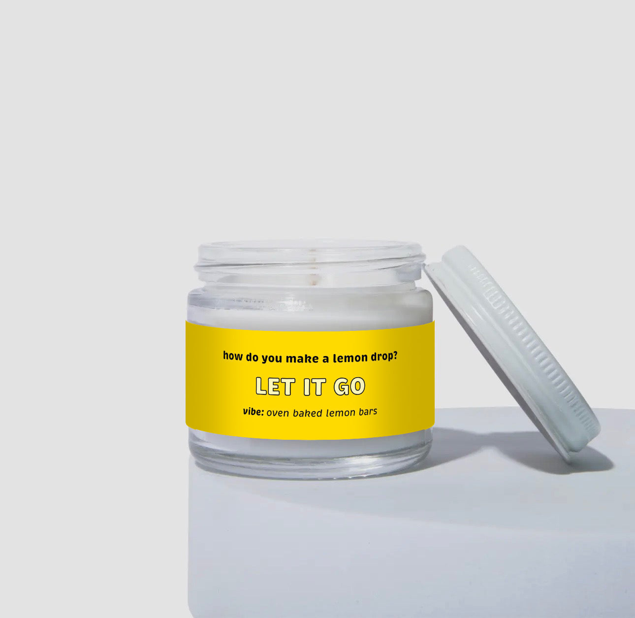 Unlit 2oz glass jar candle with a white lid, featuring a bright yellow label that reads: “How do you make a lemon drop? LET IT GO.” Scent note on the label says: “vibe: oven baked lemon bars.” Minimalist product display.