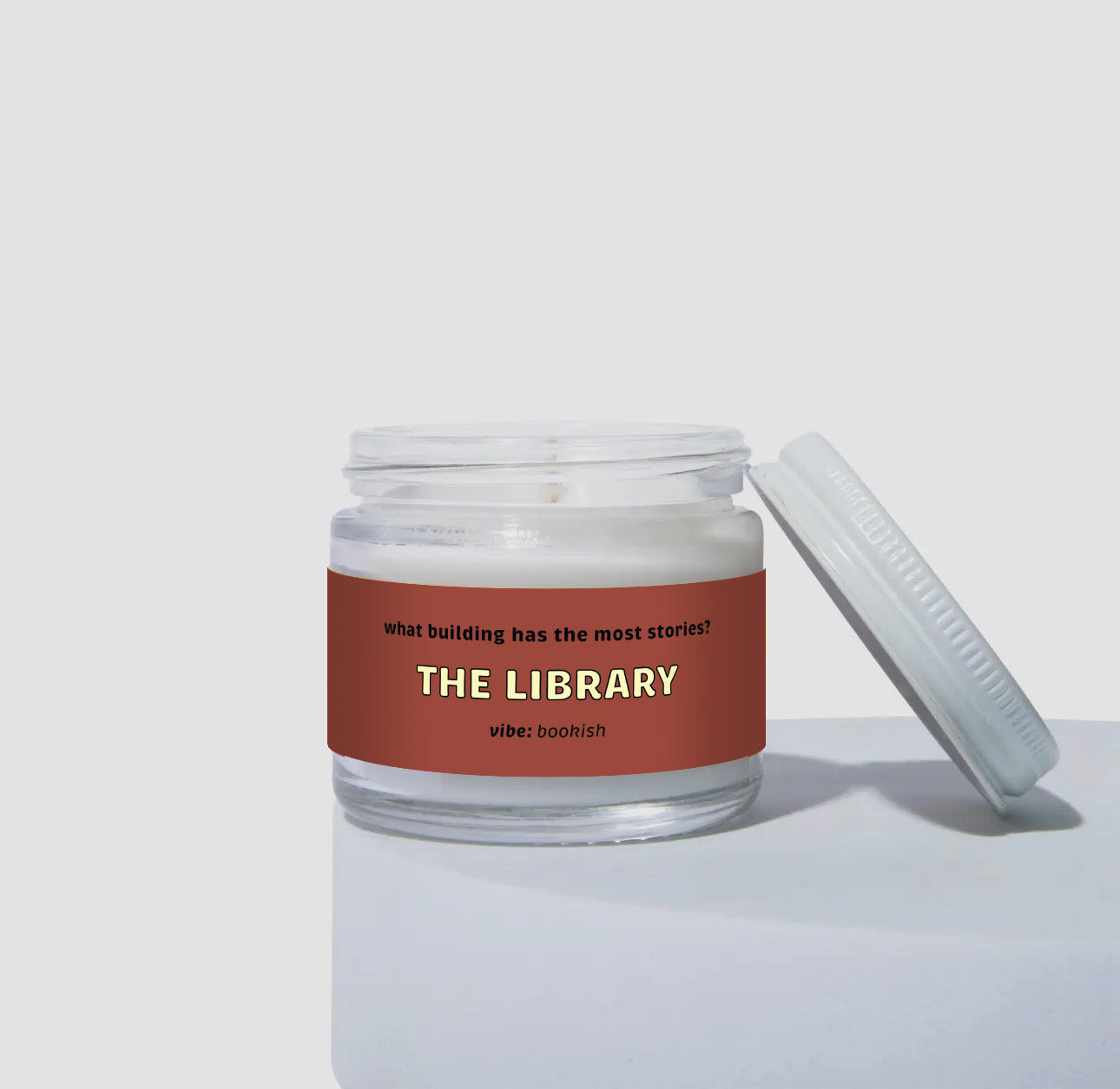 Unlit 2oz glass jar coconut soy candle with a white lid, featuring a deep red label that reads: “what building has the most stories? THE LIBRARY.” Scent note on the label says: “vibe: bookish.” Minimalist product photo against a light neutral background.