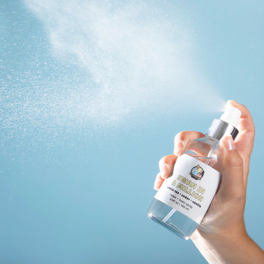 Hand holding and spraying a clear glass bottle of room + linen spray labeled 'SWAN IN A MILLION' with scent notes of white tea, cedar, and vanilla. Mist is visibly dispersing against a soft blue background, showing the fine spray in action. Bottle size: 3.4 fl oz / 100 ml.