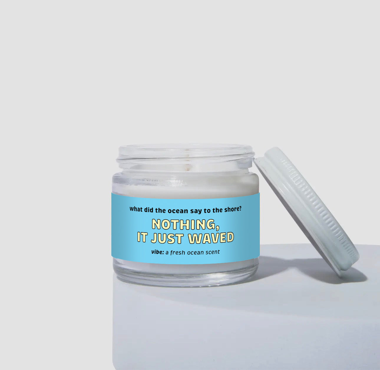 Unlit 2oz mini glass jar coconut soy candle with white lid placed to the side. The candle features a bright blue label with the text: 'what did the ocean say to the shore? NOTHING, IT JUST WAVED.' Scent description reads: 'vibe: a fresh ocean scent.' Clean product photo on a neutral background.