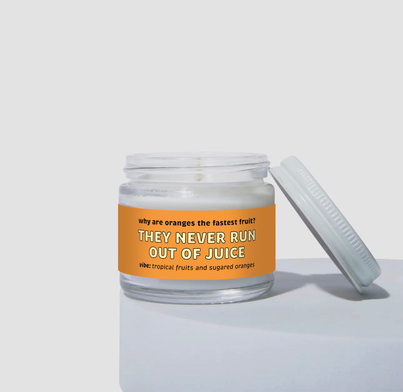 Unlit 2oz glass jar coconut soy candle with a white lid, featuring a bright orange label that reads: “Why are oranges the fastest fruit? THEY NEVER RUN OUT OF JUICE.” Scent note on the label says: “vibe: tropical fruits and sugared oranges.” Minimalist product display.