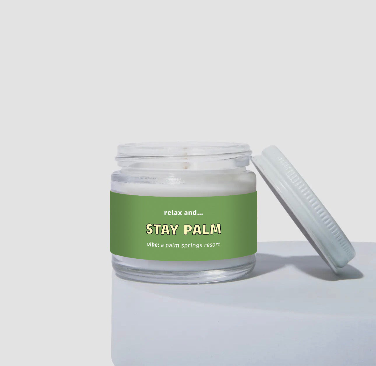 2 oz mini coconut soy candle in a clear glass jar with a white lid, labeled “Stay Palm.” Green label with the text “relax and… Stay Palm,” with the vibe described as “a Palm Springs resort.