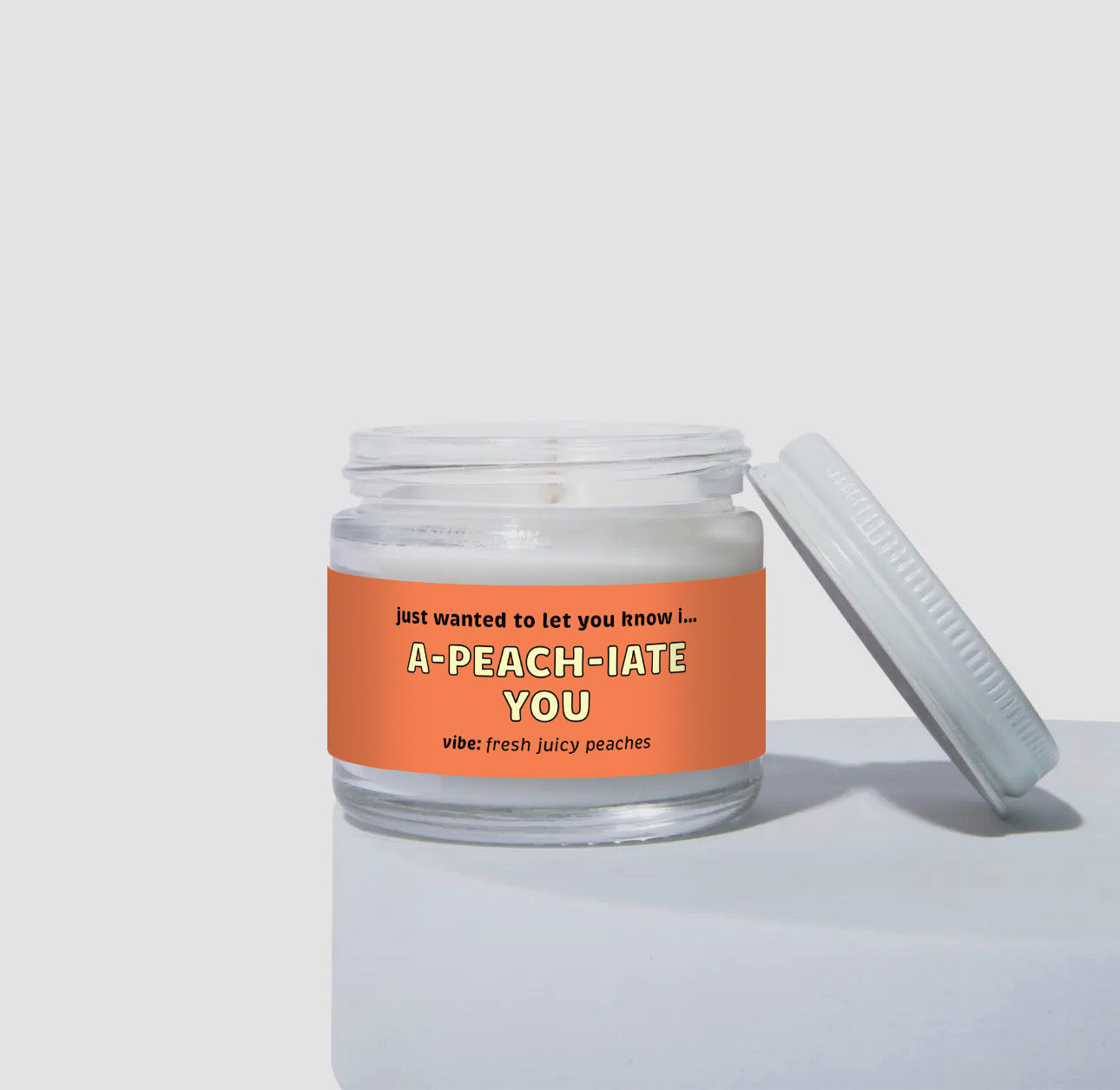 Unlit 2oz glass jar coconut soy candle with a white metal lid resting beside it. The candle features a bright peach-orange label that says: 'just wanted to let you know I… A-PEACH-IATE YOU.' Scent note reads: 'vibe: fresh juicy peaches.' Minimalist product shot highlighting the jar and lid.