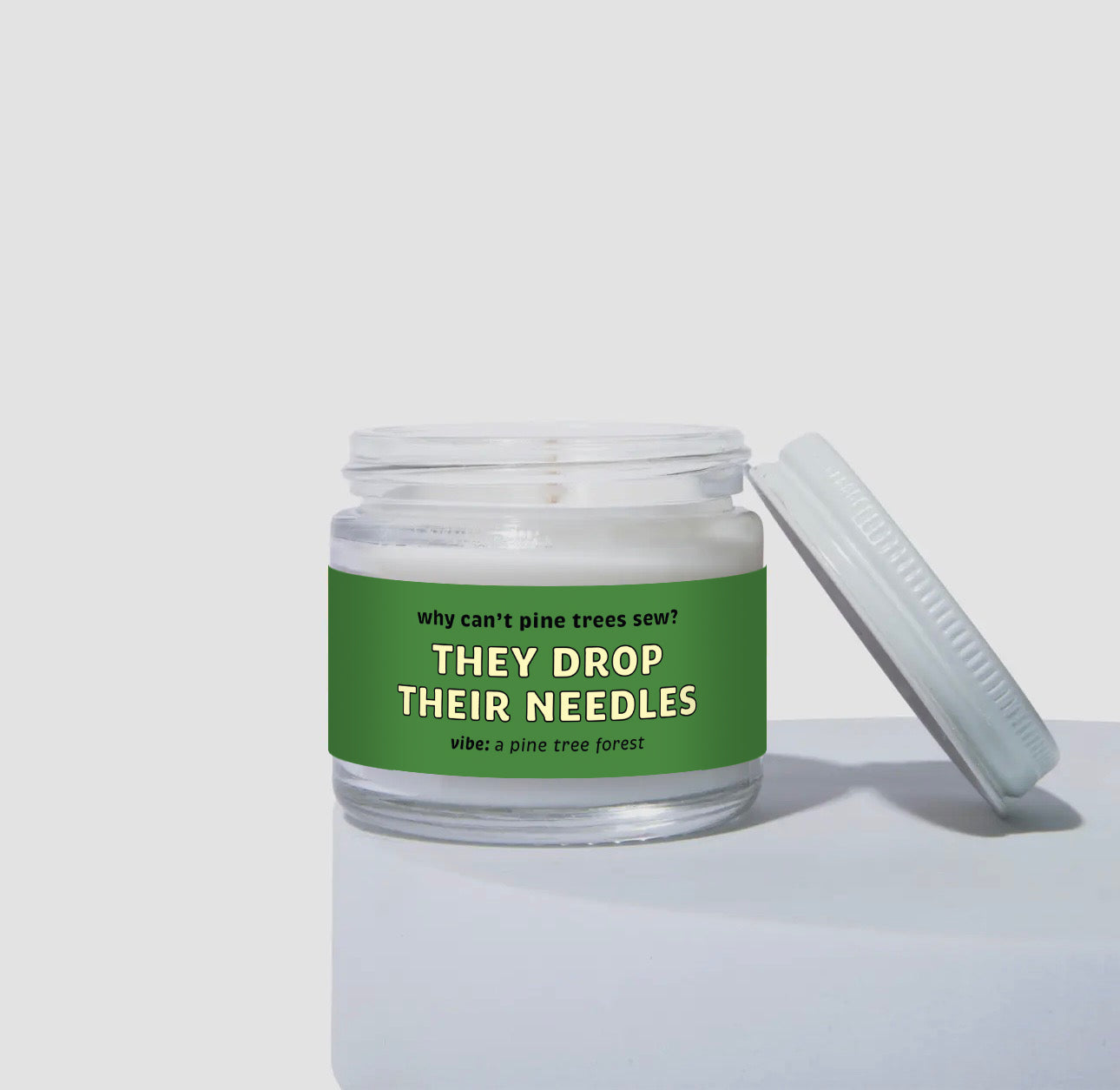 2 oz mini coconut soy candle in a clear glass jar with a white lid, labeled “They Drop Their Needles.” Green label features the text “Why can’t pine trees sew? They drop their needles” with the vibe described as “a pine tree forest.”