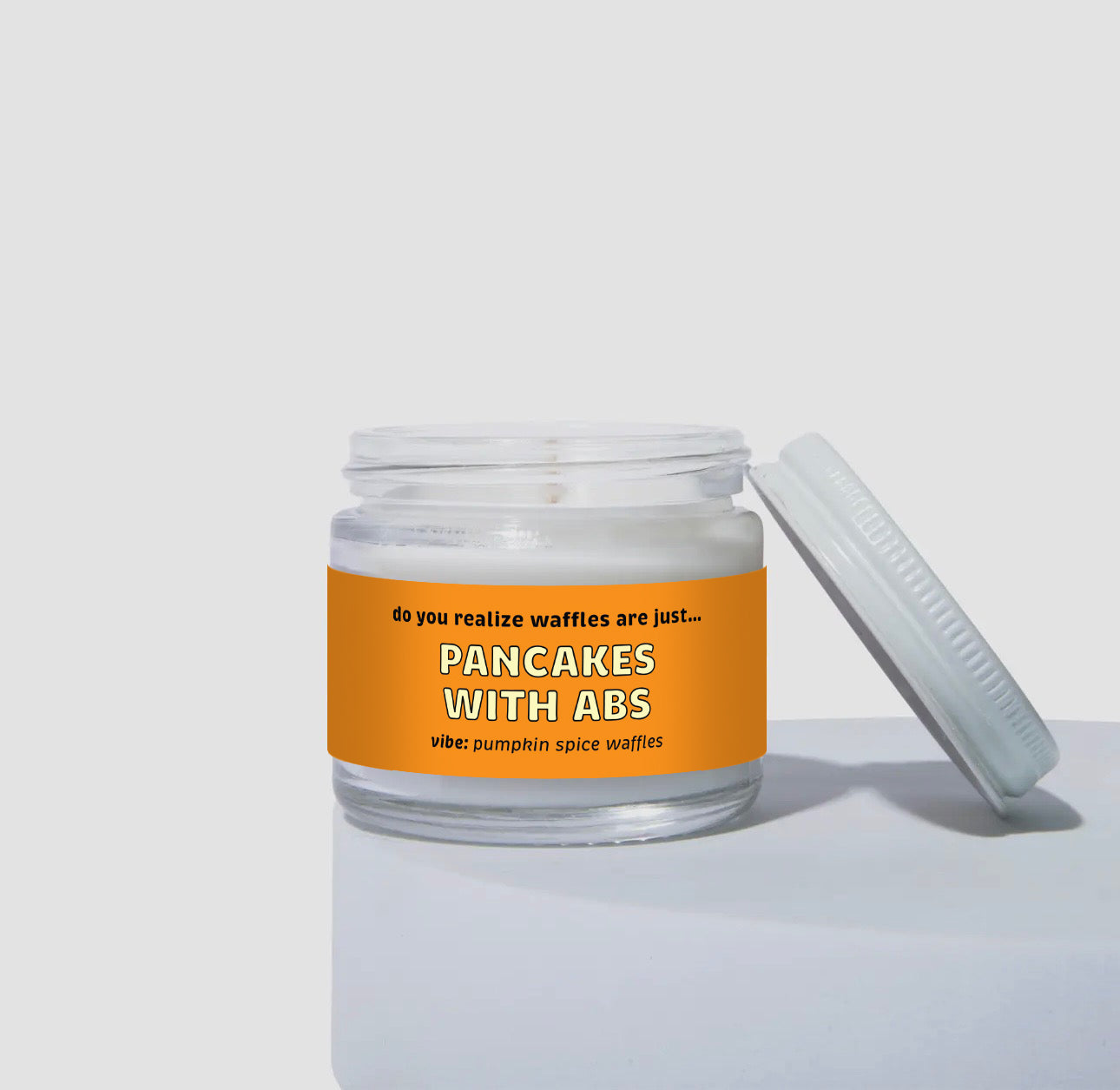Unlit 2oz glass jar coconut soy candle with a white lid, featuring a bold orange label that reads: “Do you realize waffles are just… PANCAKES WITH ABS.” Scent note on the label says: “vibe: pumpkin spice waffles.” Minimalist product photo against a neutral background.