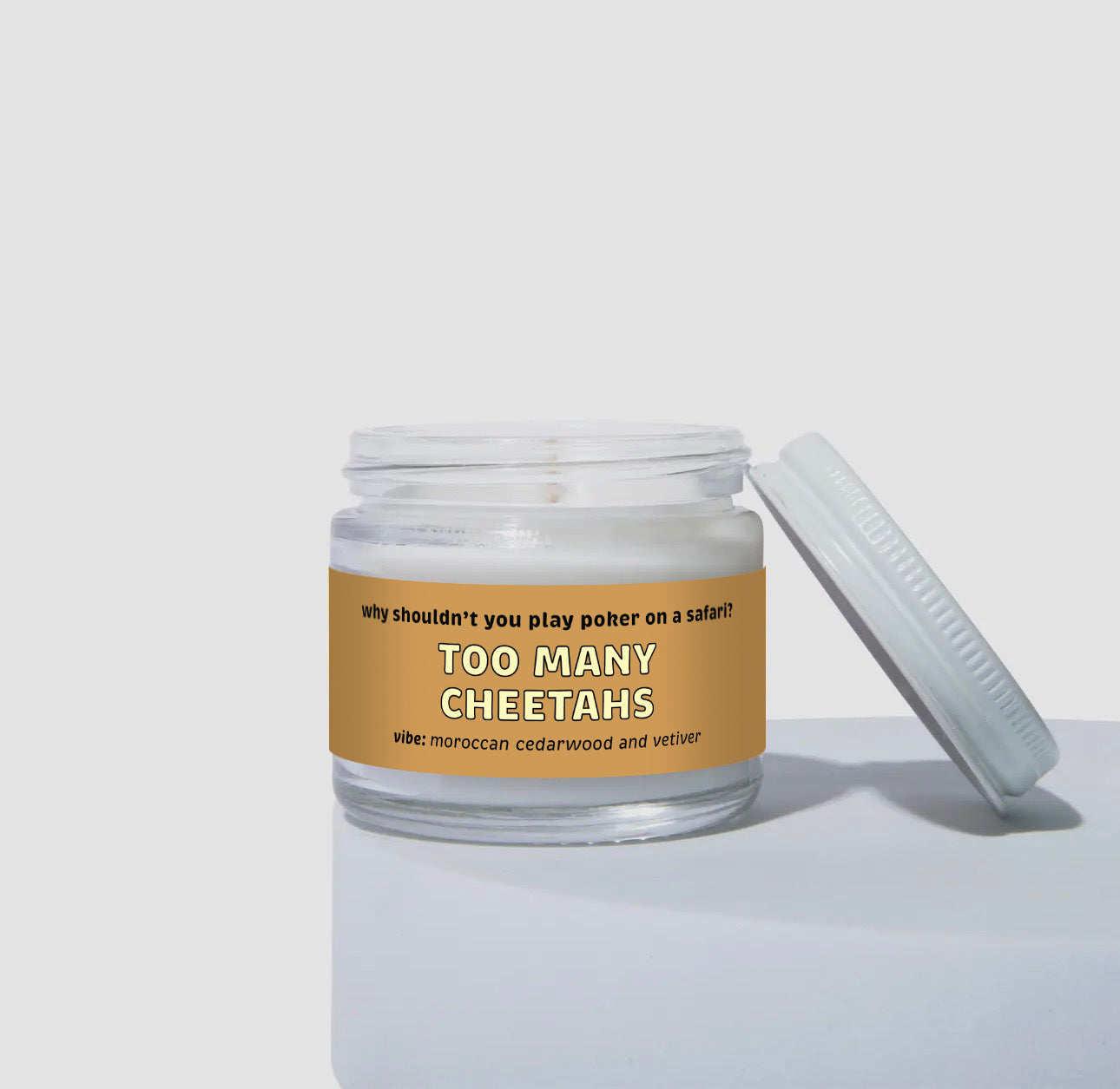 2 oz mini coconut soy candle in a clear glass jar with a golden brown label that reads “Too Many Cheetahs.” The design includes the text “why shouldn’t you play poker on a safari?” and the vibe is moroccan cedarwood and vetiver. White wax inside jar with lid resting against the side.