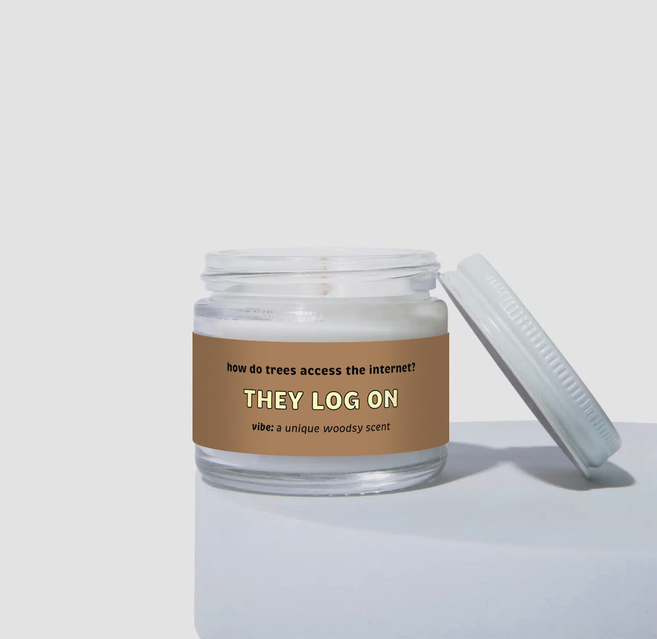Unlit 2oz glass jar coconut soy candle with a white lid, featuring a brown label that reads: “How do trees access the internet? THEY LOG ON.” Scent note on the label says: “vibe: a unique woodsy scent.” Minimalist product display.