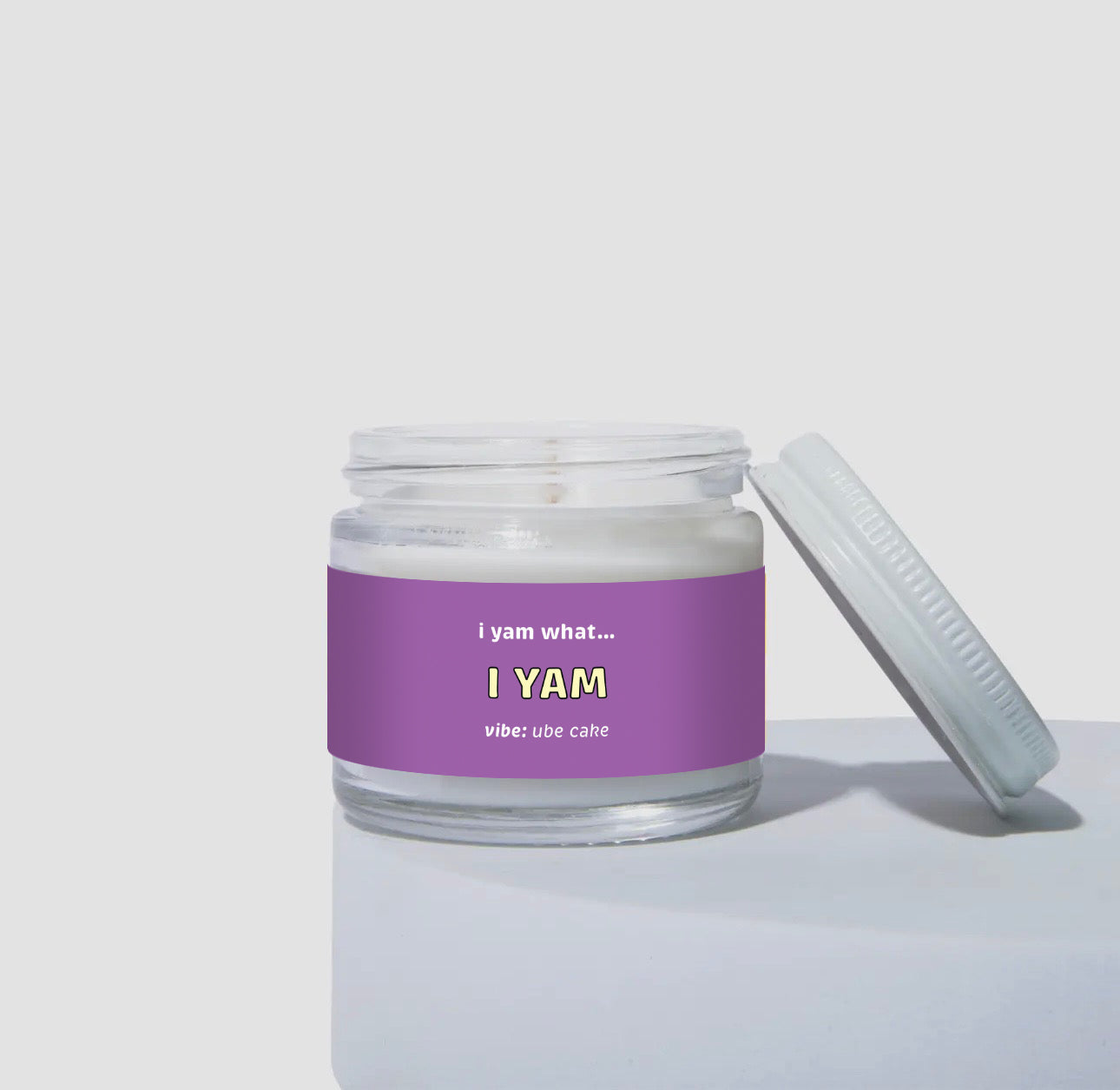 Unlit 2oz mini glass jar coconut soy candle with a white lid resting to the side. The candle features a bold purple label with the text: 'i yam what... I YAM.' Scent description reads: 'vibe: ube cake.' Clean product photo on a neutral background.