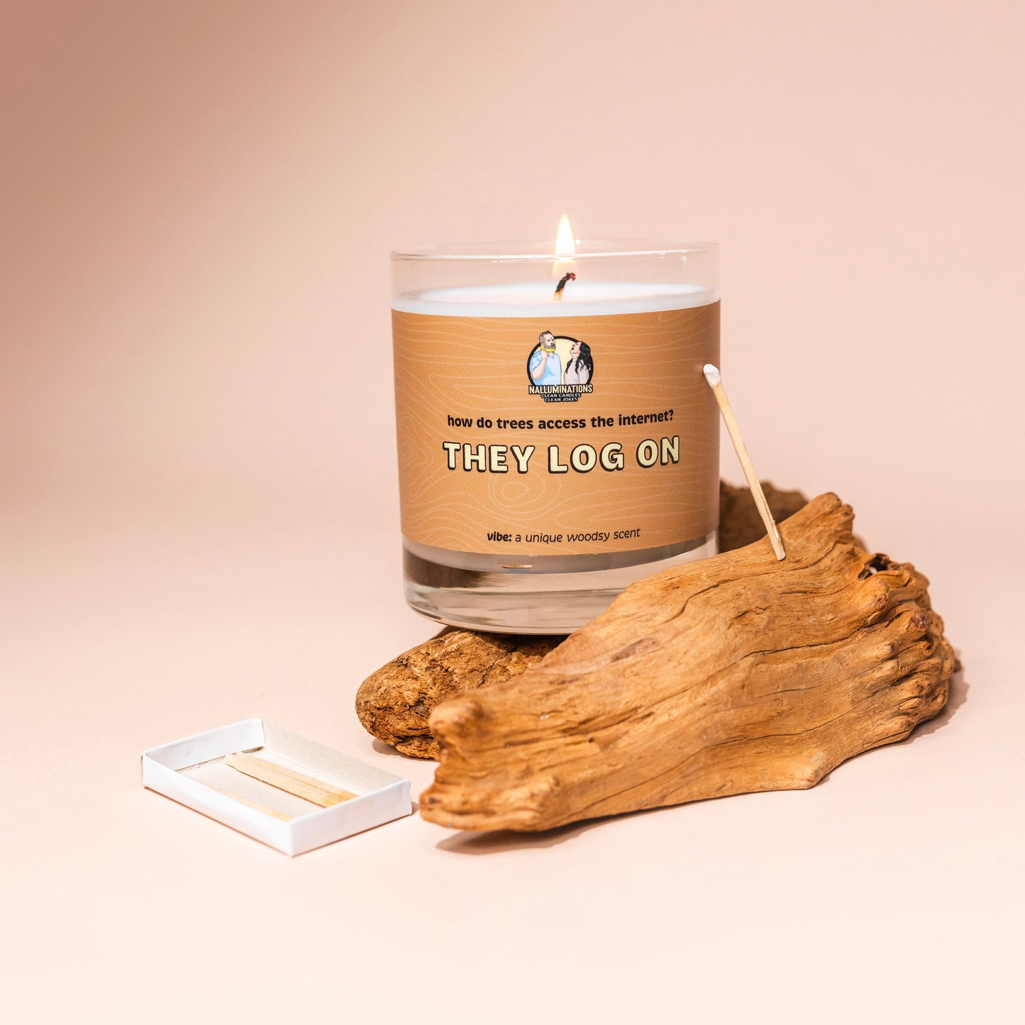 Lit 8oz glass jar candle with coconut-soy wax and a textured brown label that says: 'how do trees access the internet? THEY LOG ON.' Scent note reads: 'vibe: a unique woodsy scent.' The candle is styled on a neutral background with driftwood, a matchstick, and an open matchbox for a rustic, natural look