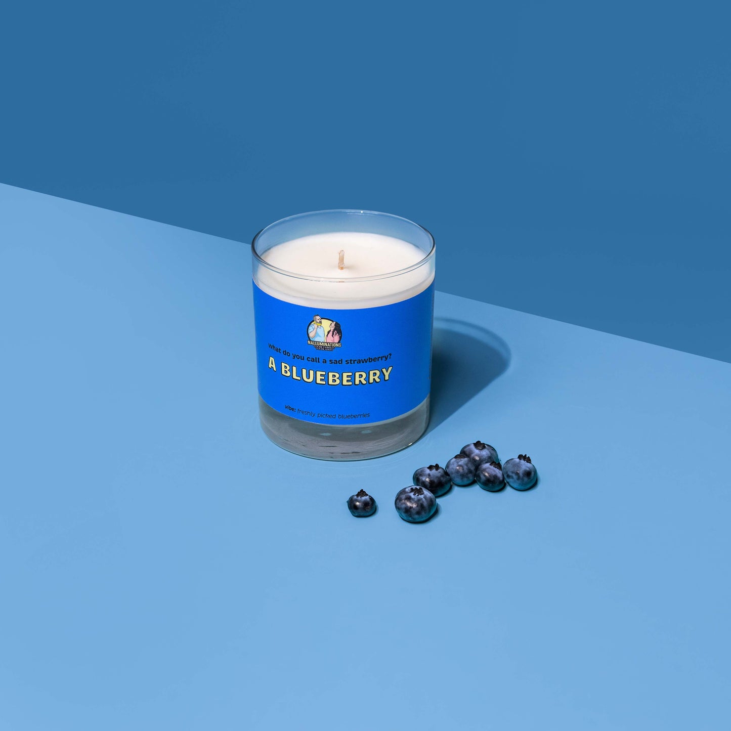 White coconut soy candle in a clear glass jar with a bright blue label that reads “A Blueberry” with the question “What do you call a sad strawberry?” above it. The scent vibe is “freshly picked blueberries.” The candle is styled on a two-tone blue background with fresh blueberries scattered in front.