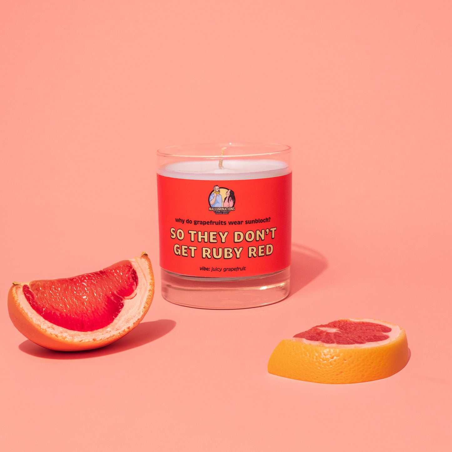 Unlit 8oz glass jar candle with coconut-soy wax and a bright red label that says: 'why do grapefruits wear sunblock? SO THEY DON'T GET RUBY RED.' Scent note reads: 'vibe: juicy grapefruit.' The candle is styled on a coral background with fresh grapefruit slices placed around it for a vibrant, citrusy look.