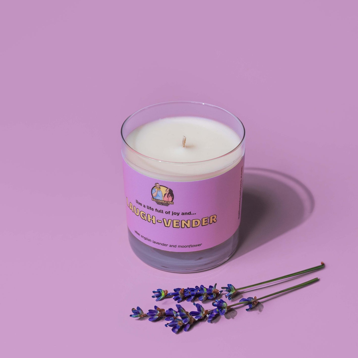 Lavender-scented coconut soy candle in a clear glass jar with a purple label reading “Laugh-Vender,” placed on a lavender-colored background with sprigs of fresh lavender in front. The vibe is described as “english lavender and moonflower.
