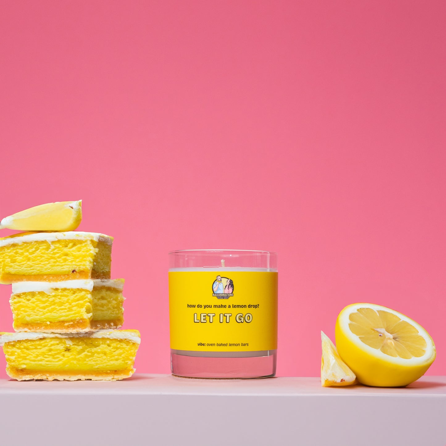 A coconut soy candle in a clear glass jar with a bright yellow label that reads “Let It Go – Oven Baked Lemon Bars.” The candle is displayed against a vibrant pink background, styled with a stack of lemon bars on the left and a halved lemon on the right.