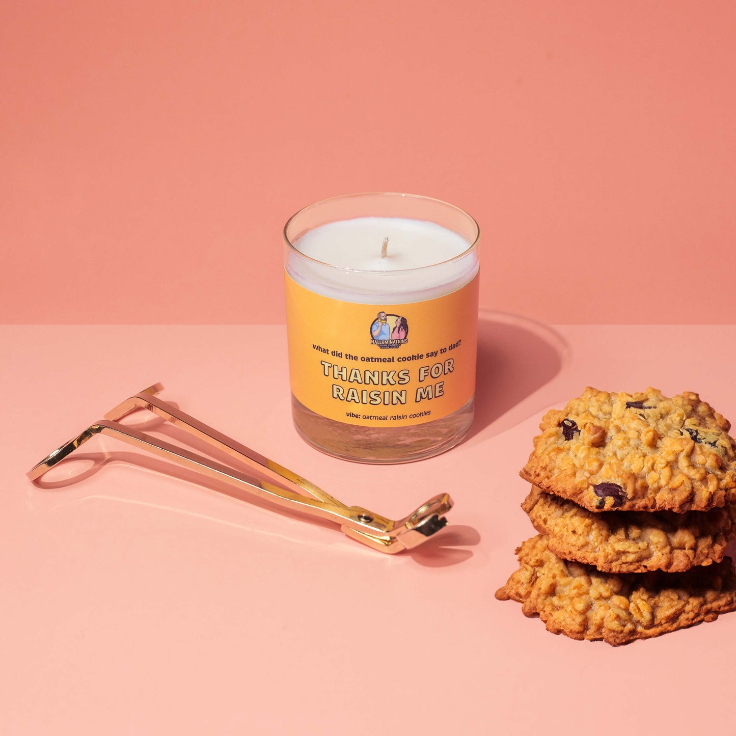 Unlit 8oz glass jar coconut soy candle with a light tan label that says: 'What did the oatmeal cookie say to dad? THANKS FOR RAISIN ME.' Scent note reads: 'vibe: oatmeal raisin cookies.' The candle is displayed on a peach-colored background next to a gold wick trimmer and a small stack of oatmeal raisin cookies.
