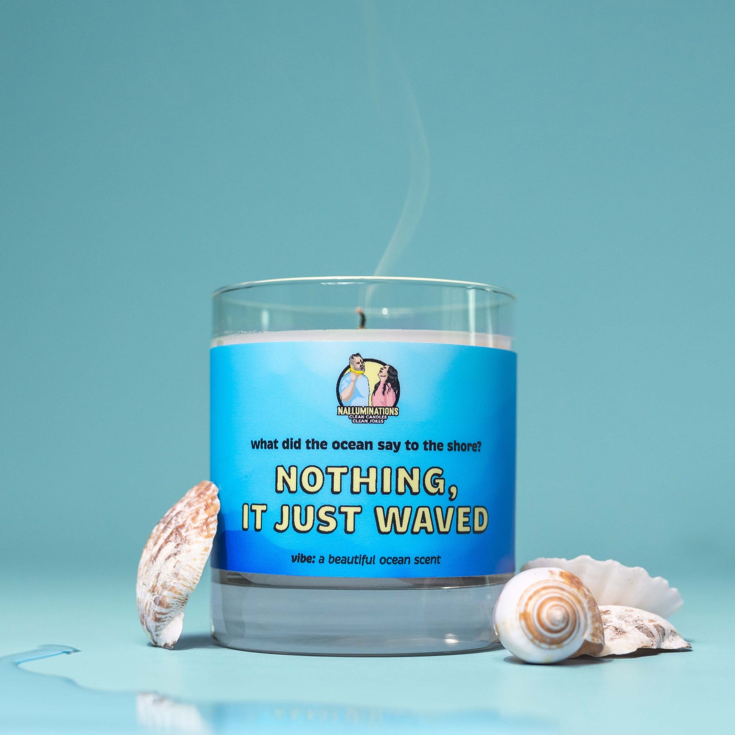 Coconut soy candle in a clear glass jar with a blue label that reads “Nothing, It Just Waved.” Fresh ocean scent, styled on a blue background with seashells around the candle.
