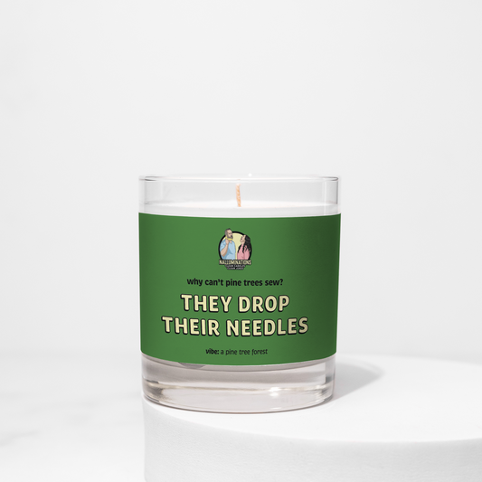 8oz coconut soy candle in a clear glass jar with a green label. Label says, “Why can’t pine trees sew? They drop their needles.” Pine tree forest fragrance, shown on a white background.