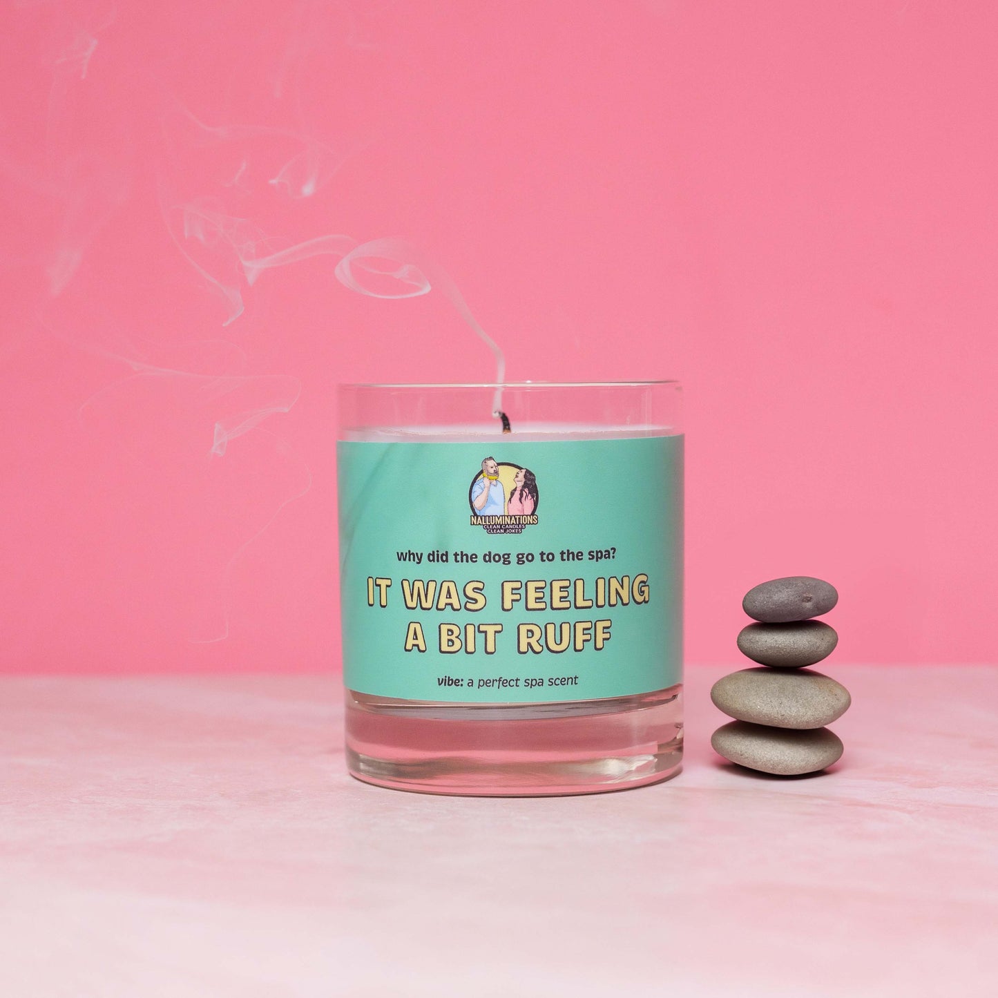 Recently extinguished 8oz glass jar candle with coconut-soy wax and a teal label that says: 'why did the dog go to the spa? IT WAS FEELING A BIT RUFF.' Scent note reads: 'vibe: a perfect spa scent.' Wisps of smoke rise from the wick as the candle sits against a pink background, styled beside a neat stack of smooth spa stones.