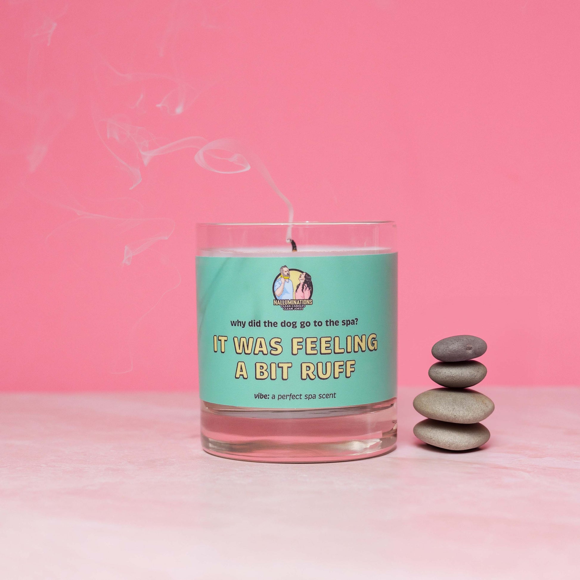 Recently extinguished 8oz glass jar candle with coconut-soy wax and a teal label that says: 'why did the dog go to the spa? IT WAS FEELING A BIT RUFF.' Scent note reads: 'vibe: a perfect spa scent.' Wisps of smoke rise from the wick as the candle sits against a pink background, styled beside a neat stack of smooth spa stones.