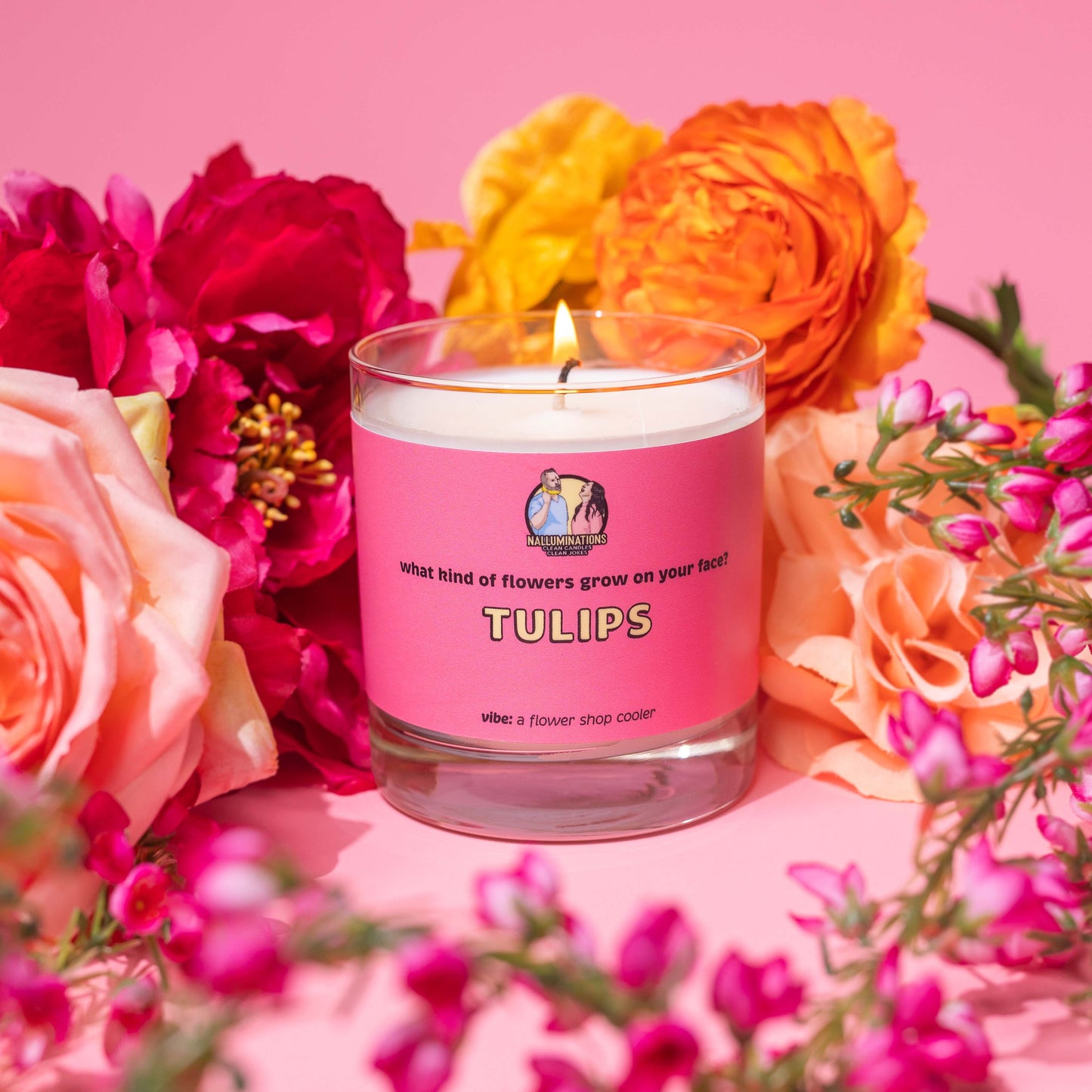 Lit 8oz glass jar candle with coconut-soy wax and a bright pink label that says: 'what kind of flowers grow on your face? TULIPS.' Scent note reads: 'vibe: a flower shop cooler.' The candle is burning, styled in a lush floral arrangement of roses, peonies, and colorful blossoms against a pink background.