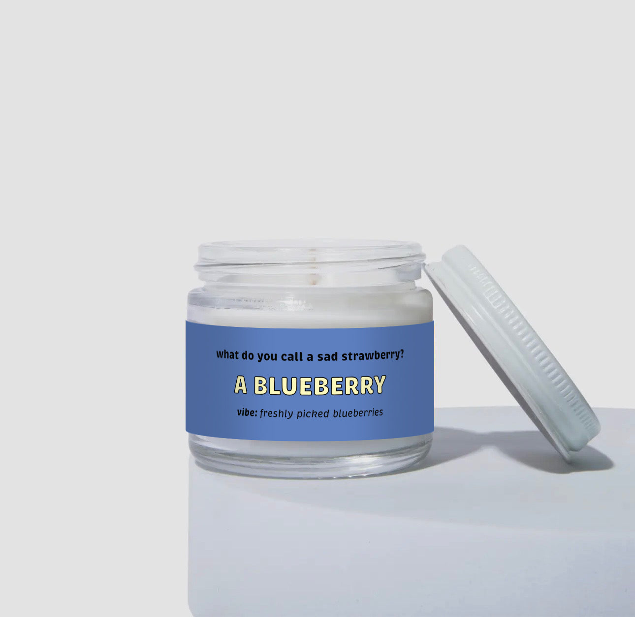 Unlit 2oz mini glass jar coconut soy  candle with a white metal lid placed to the side. The candle has a deep blue label with the text: 'What do you call a sad strawberry? A BLUEBERRY.' Scent description reads: 'vibe: freshly picked blueberries.' Clean and simple product display.