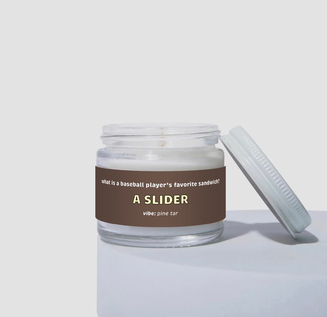 Unlit 2oz glass jar coconut soy candle with a white lid, featuring a dark brown label that reads: “what is a baseball player’s favorite sandwich? A SLIDER.” Scent note on the label says: “vibe: pine tar.” Minimalist product photo against a light neutral background.