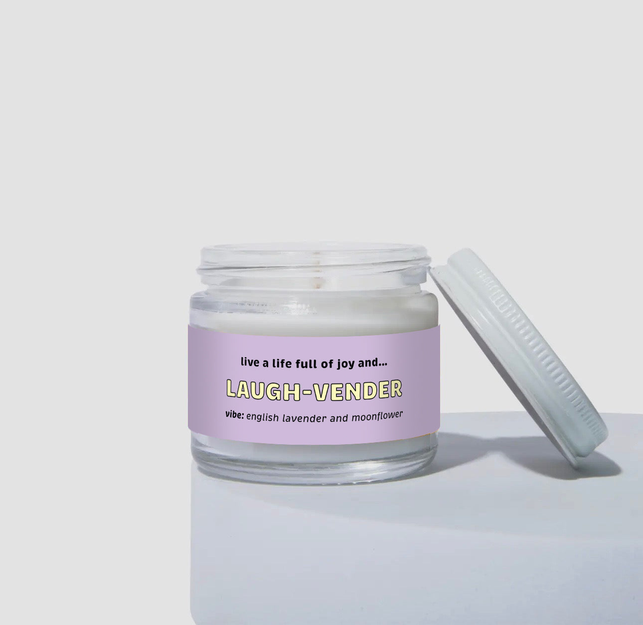 Unlit 2oz mini glass jar coconut soy candle with a white lid leaning to the side. The candle features a soft lavender-colored label that says: 'Live a life full of joy and… LAUGH-VENDER.' Scent description reads: 'vibe: English lavender and moonflower.' Simple studio product shot.