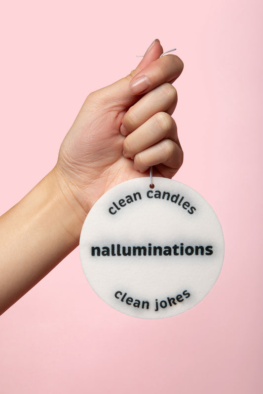 A hand holding a round white novelty candle tag with black text reading 'clean candles', 'nalluminations', and 'clean jokes' against a solid pink background.