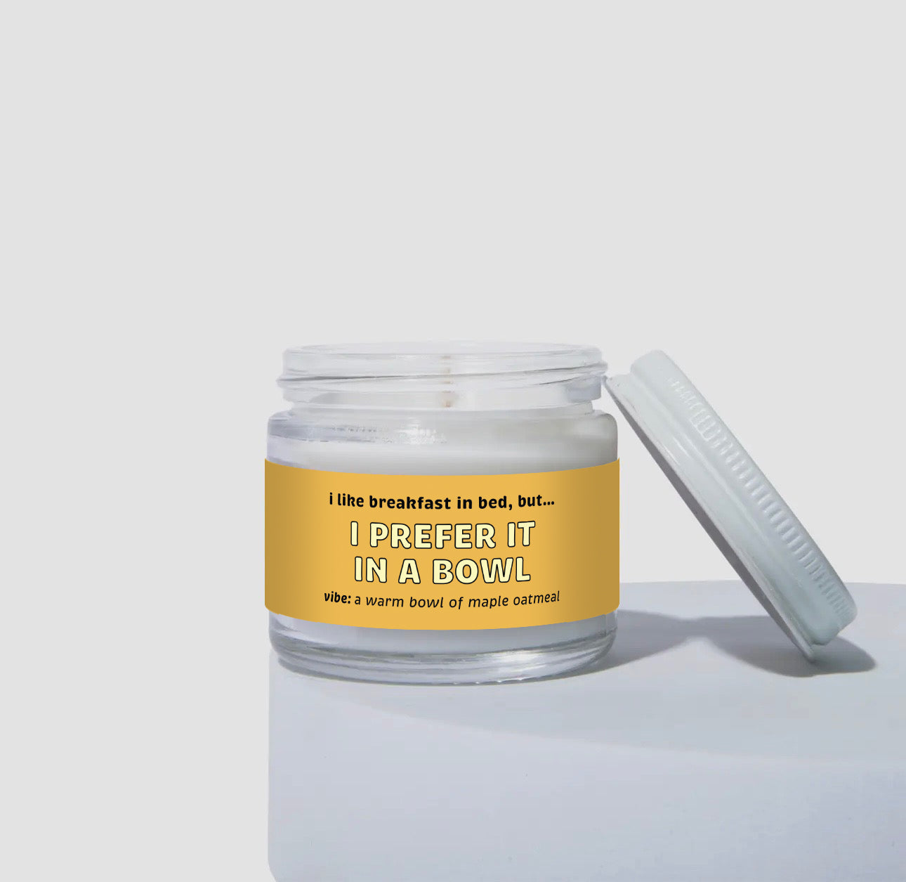 Unlit 2oz mini glass jar coconut soy candle with a white metal lid resting beside it. The candle features a warm golden-yellow label that says: 'I like breakfast in bed, but… I PREFER IT IN A BOWL.' Scent note reads: 'vibe: a warm bowl of maple oatmeal.' Clean, minimalist product display.