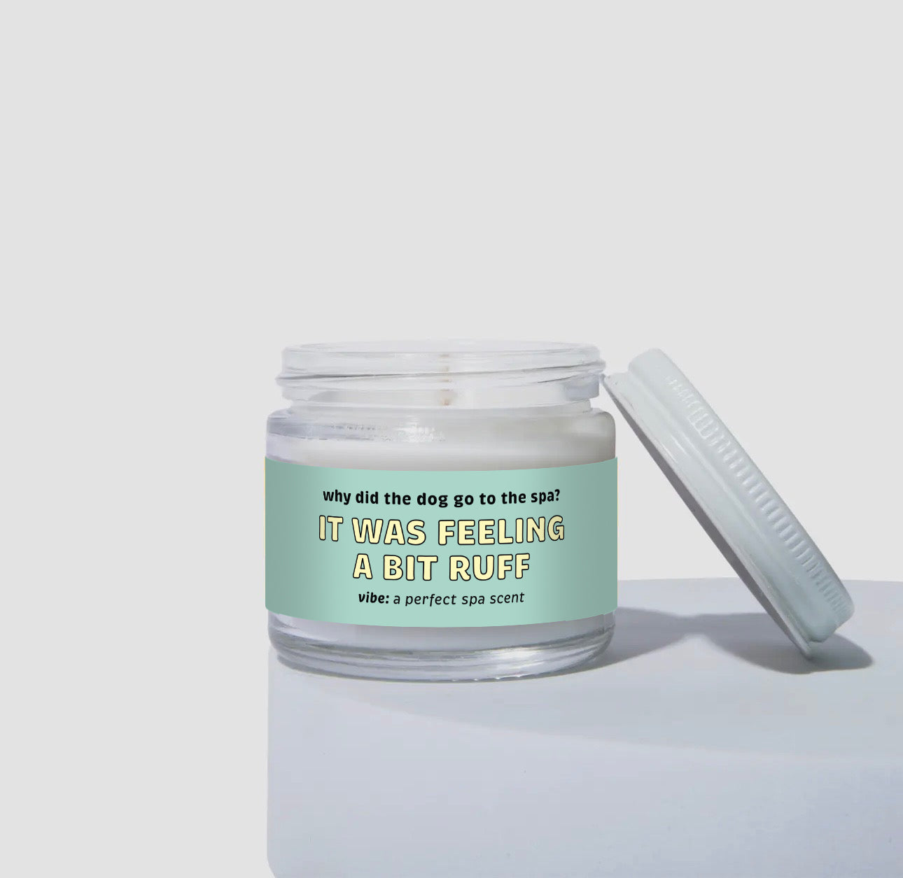 Unlit 2oz glass jar candle with a white lid, featuring a pastel aqua-green label that reads: “Why did the dog go to the spa? IT WAS FEELING A BIT RUFF.” Scent note on the label says: “vibe: a perfect spa scent.” Minimalist product photo against a neutral background.